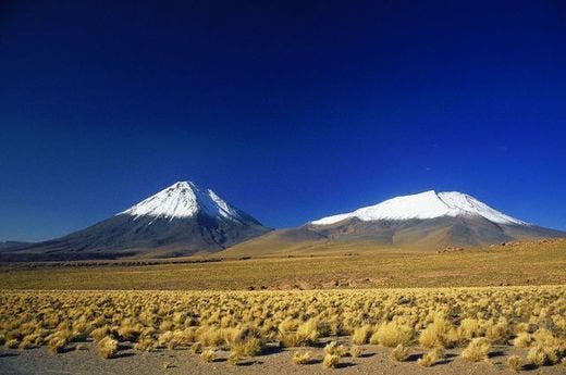 White-peaked volcanos rise from the plains of the Atacama Desert under deep blue skies. The Atacama is the driest desert in the world.