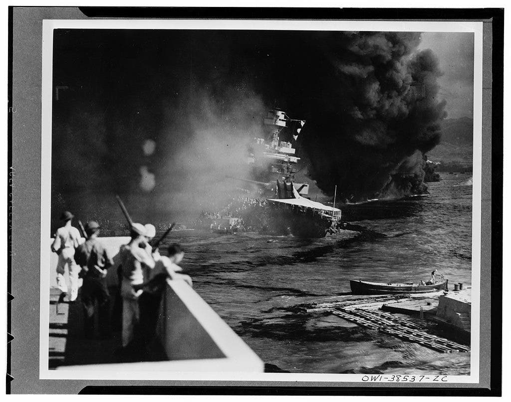California hit. Battered by aerial bombs and torpedoes, the U.S.S. California settles slowly into the mud and muck of Pearl Harbor. Clouds of black oily smoke pouring up from the California and her stricken sister ships conceal all but the hulk of the capsized U.S.S. Oklahoma at extreme right. December 1941