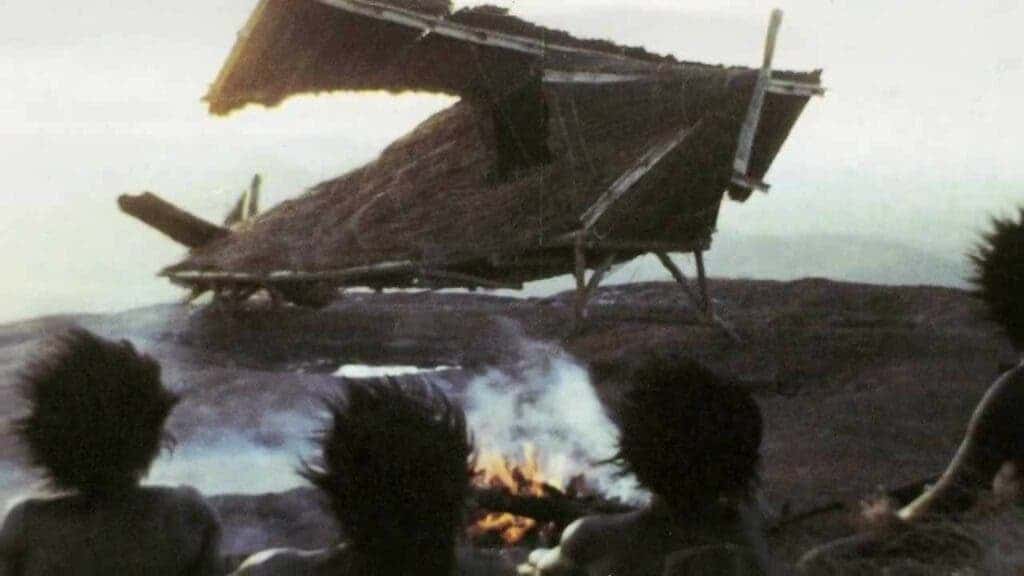 Photo of cargo cult people gathered around fire with straw airplane in background. 