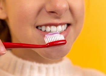 A girl smiling and holding a toothbrush (with toothpaste on it) in her hands.